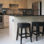 ottawa modern kitchen renovations Modern kitchen renovation with grey floor tile, off-white painted walls, black countertops, natural wood cupboards, and a black, white and grey tile patterned backsplash.