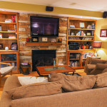 basement renovation ottawa Renovated basement with hardwood floors, yellow painted walls, wood bookshelves, windowed fireplace surrounded by a stone facade with a TV mounted above and ceiling mounted speakers.